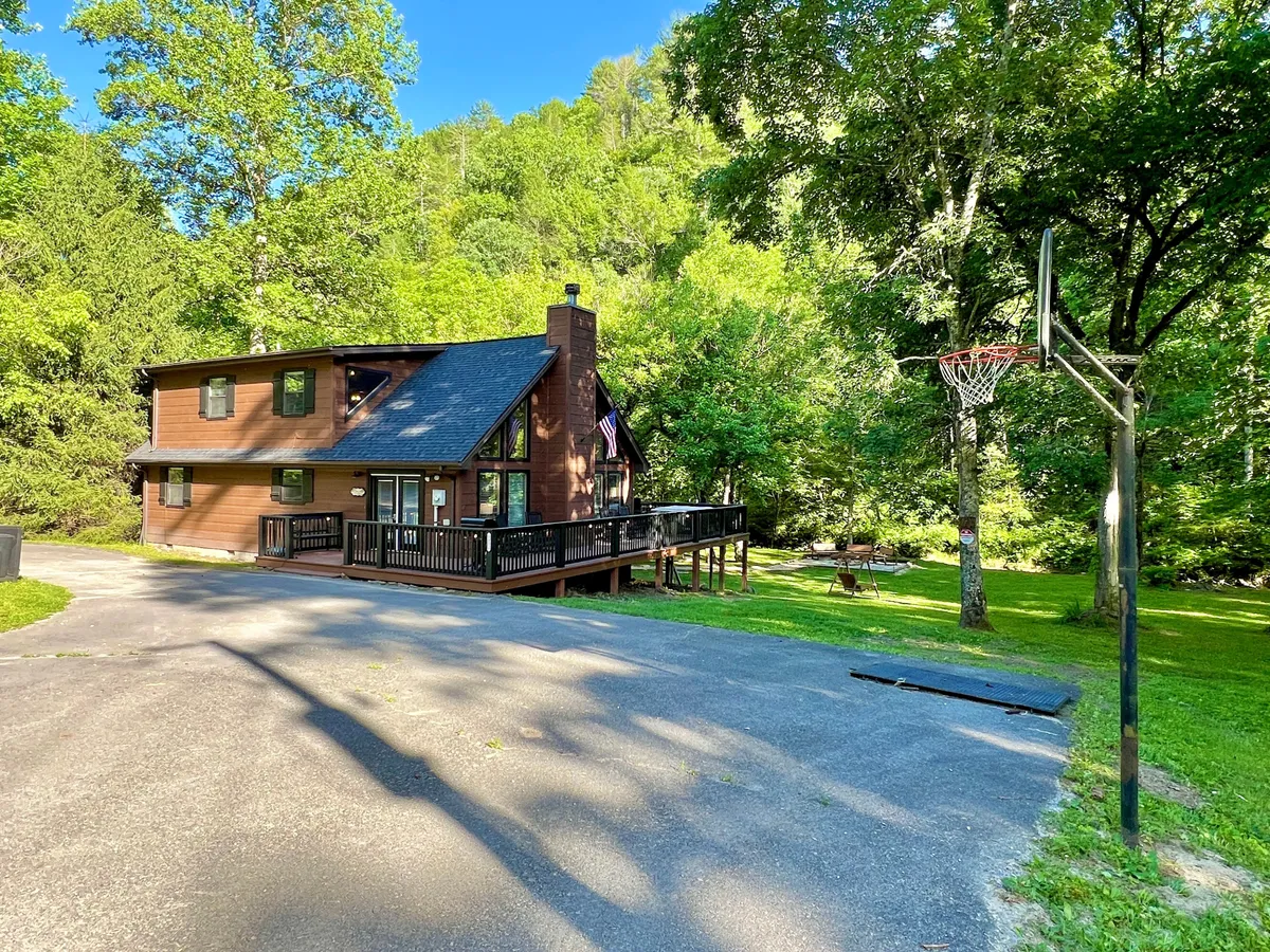 The front of the house is shown with a basketball hoop, an expansive deck, and mature trees in the background. The area is open and pet-friendly, ideal for both lively playtimes and peaceful relaxation.