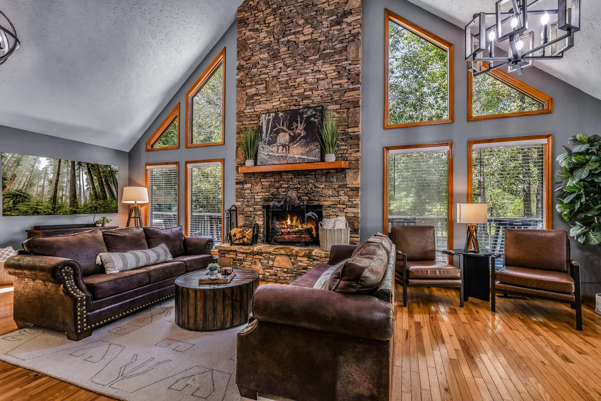 A cozy living room setting with a beautiful stone fireplace, large windows that showcase woodland views, comfortable sofas and armchairs, and a wooden coffee table. Warm natural light fills the room, and the arrangement invites relaxation or conversation.