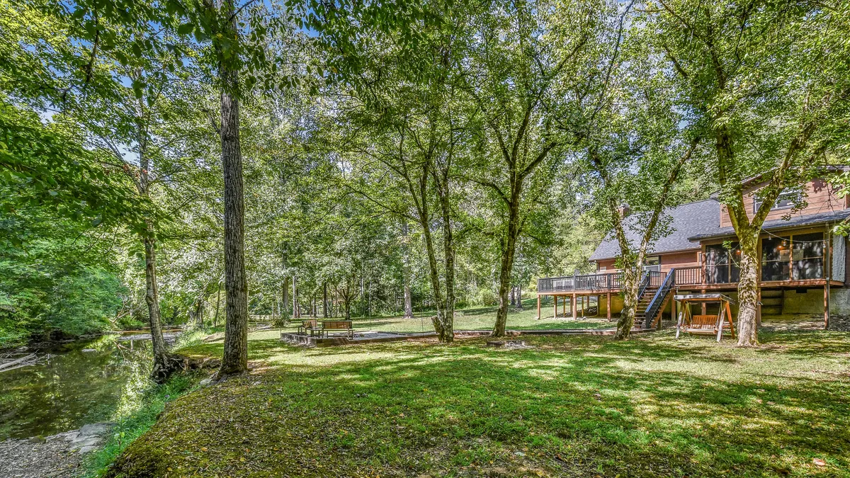 A peaceful exterior view featuring a lush riverbank, outdoor seating area, and the deck of the house. The scene is set against vibrant greenery with the tranquil water nearby, suggesting close proximity to nature and easy access to outdoor activities.