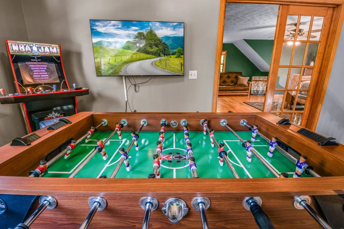 The scene presents a lively corner of the game loft with a foosball table, a TV displaying scenic views, and an arcade game machine. The space feels energetic and inviting, ideal for gaming or relaxing after a day of adventure.