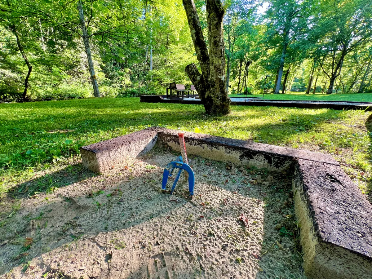 An exterior play area featuring a sandbox with a blue rake, surrounded by green grass and tall trees, offering a classic, inviting space for children to play and families to relax outdoors.