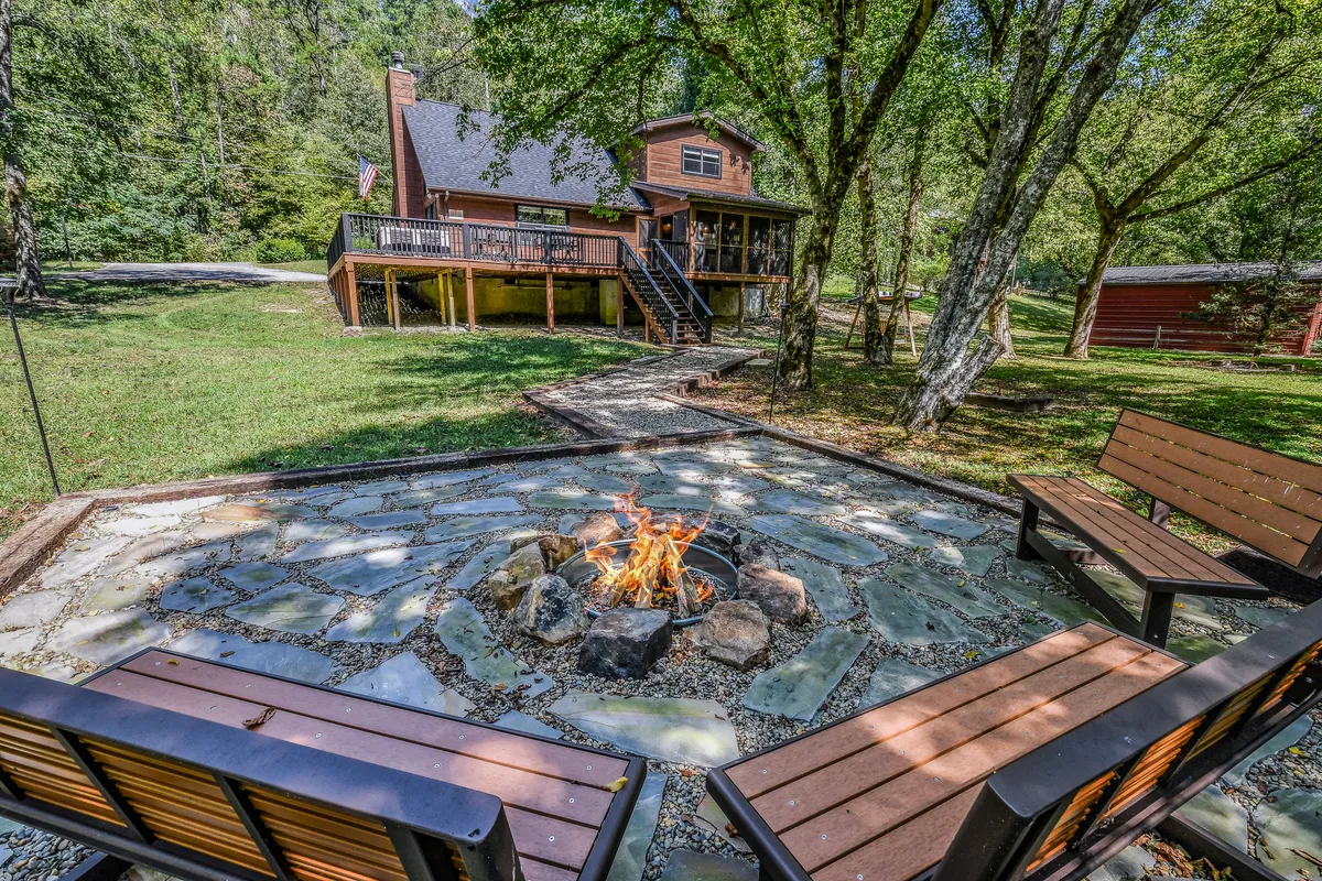 A cozy fire pit area with wooden benches is set against a backdrop of trees and the house deck, inviting guests to gather, share stories, and enjoy evenings by the fire under the stars.