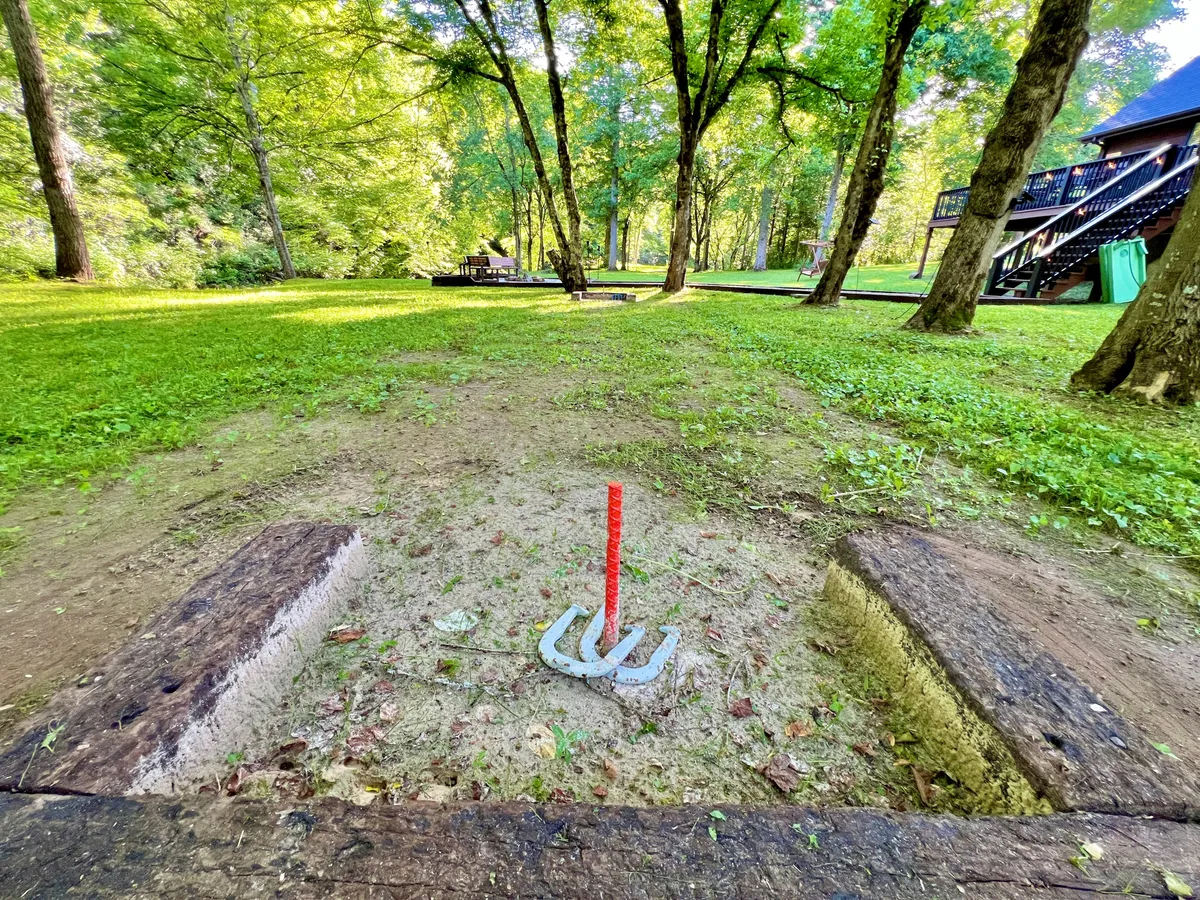A horseshoe pit sits amid lush green grass and shade trees, with a picnic area and the home’s deck visible in the background. The spacious, flat yard welcomes outdoor games and family fun.