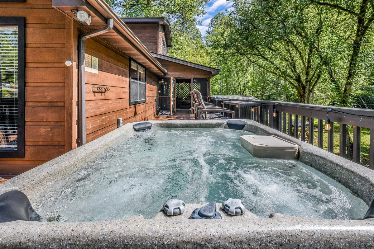 The hot tub is featured on a wooden deck surrounded by trees, creating an inviting spot for relaxation and socializing in nature. The serene setting is perfect for stargazing or enjoying a morning coffee, illustrating the cabin's outdoor amenities.
