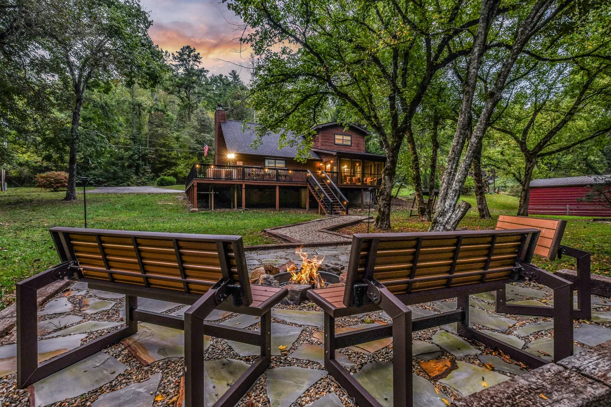 The image shows a cozy fire pit surrounded by wooden chairs, set against the backdrop of the house exterior and a lush stand of trees. The arrangement suggests a welcoming atmosphere for guests and pets to gather, relax, and unwind after a day outdoors, with plenty of space and nature all around.