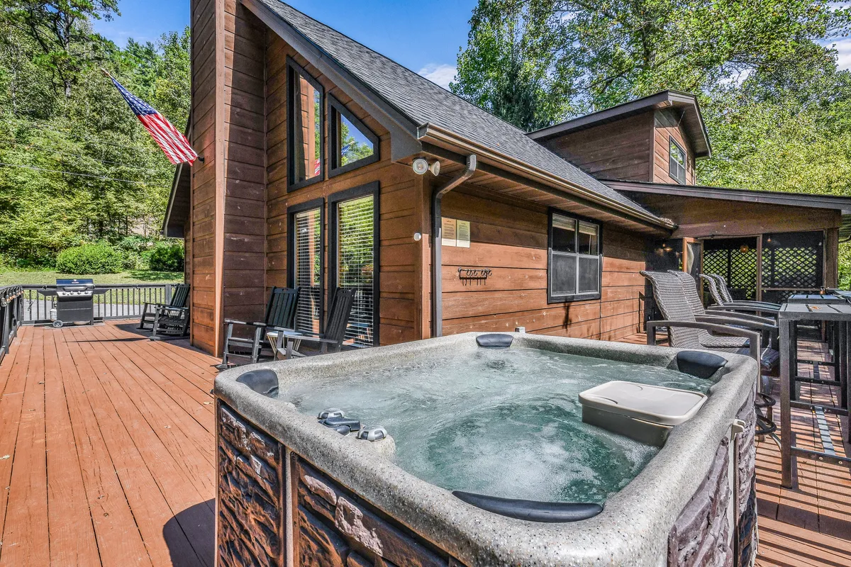A welcoming deck area featuring a bubbling hot tub, an American flag, and views of surrounding trees. The space invites relaxation and togetherness, perfect for guests to unwind after a day of local exploration.